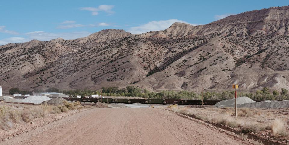 A picture of a road leading to a processing plant with a train in front of it. In the background is a mountain.