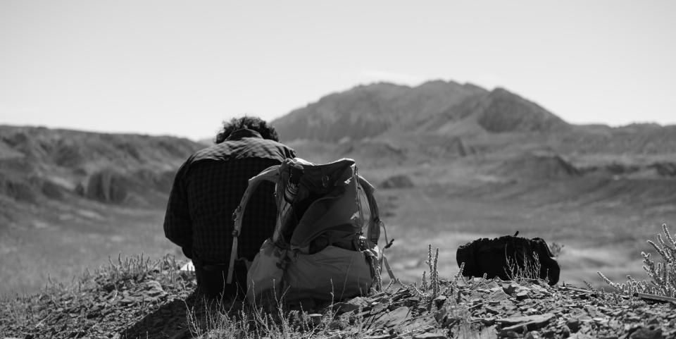 Black and white photo of a man writing notes with his back to the camera. In the distant foreground is a mountain.