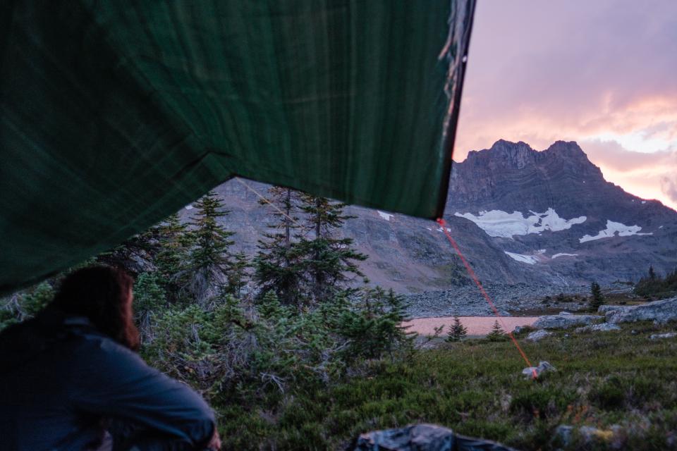 Man peering out from under a tarp. He is looking at a mountain with a pink sky behind it.